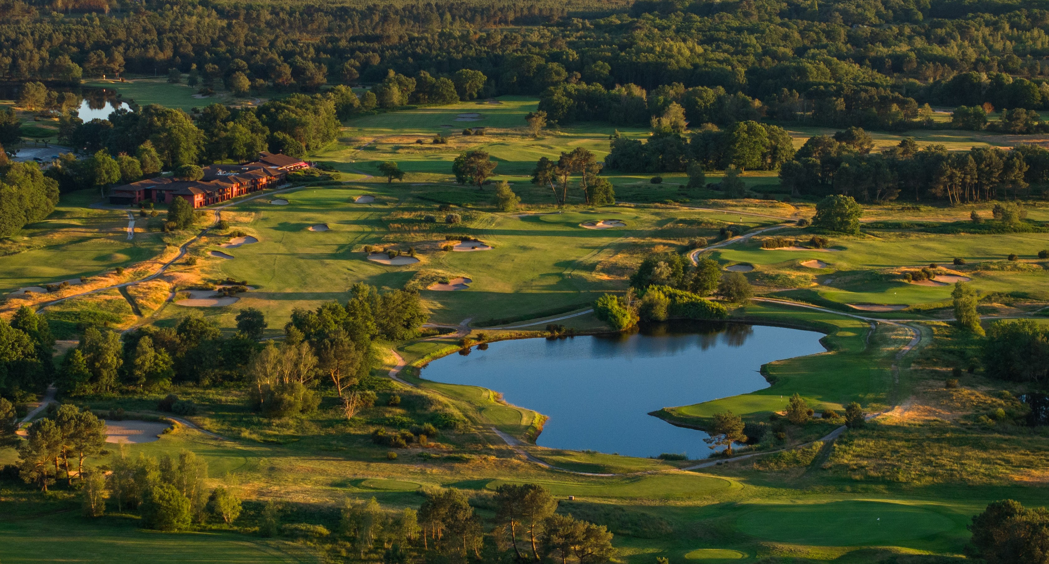 Vue aérienne du domaine Cabot Bordeaux en lumière dorée&nbsp;: l'hôtel, les fairways, les bunkers et un lac entourés de pins