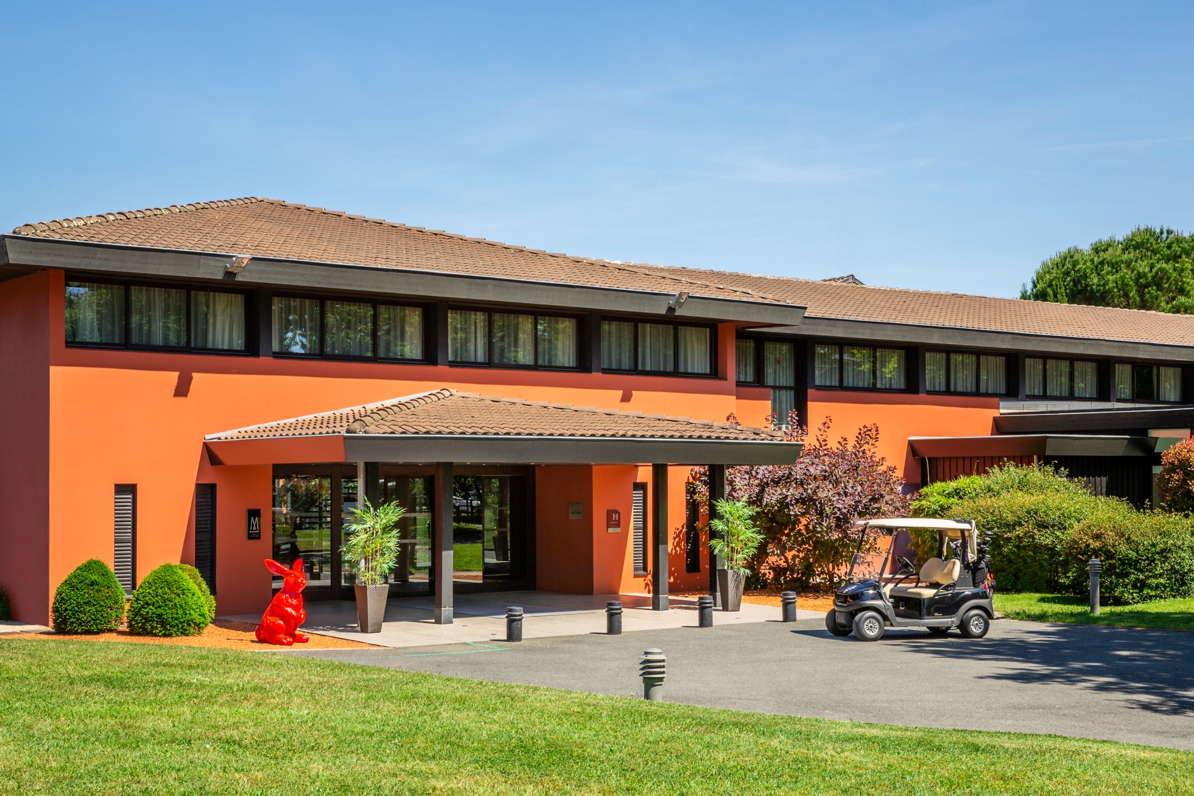 Façade de l'hôtel Cabot Bordeaux MGallery au Pian-Médoc, entrée sous auvent, pelouse et voiturette de golf sous un ciel bleu
